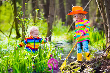 Zwei Kinder in farbenfroher Regenkleidung erkunden einen seichten Bach in einem üppigen Wald, wobei das eine ein rosa Netz und das andere einen gelben Stock hält.